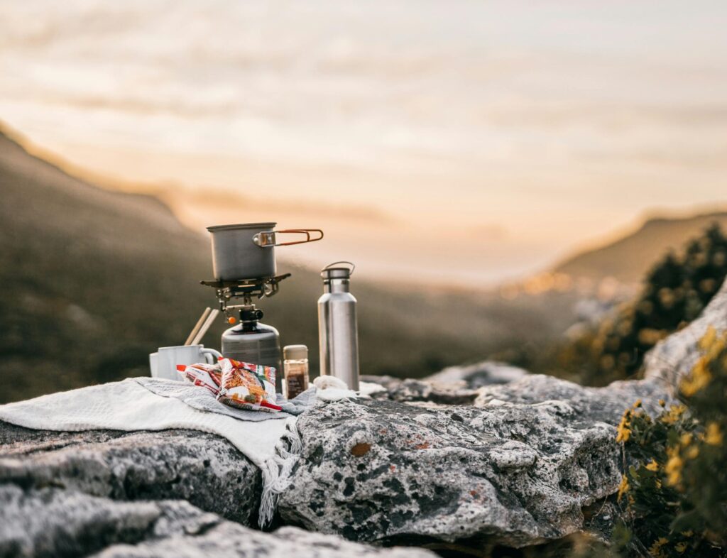 pexels-photo-9508453-9508453 A scenic outdoor camping setup with cooking gear on rocky terrain during sunrise in a mountain landscape.